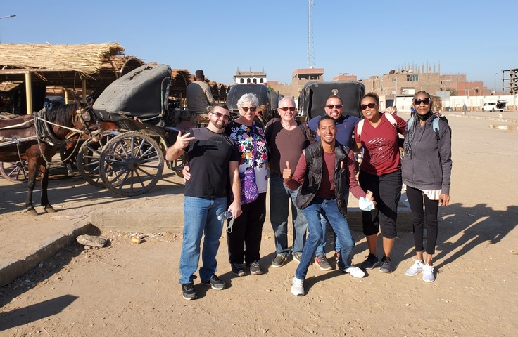       Group of tourists standing in a sunny area with carriages and buildings.
  