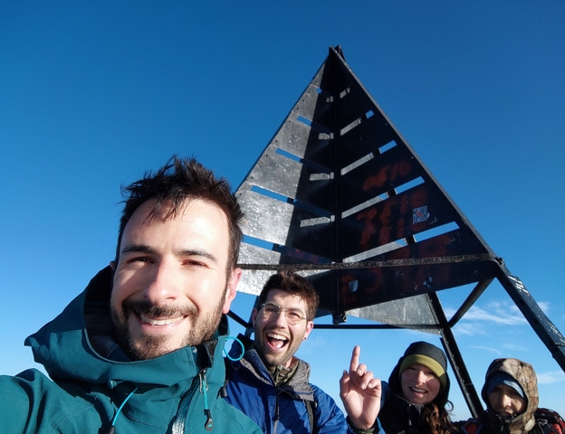 Two people smiling at the summit of a mountain with triangular monument.
