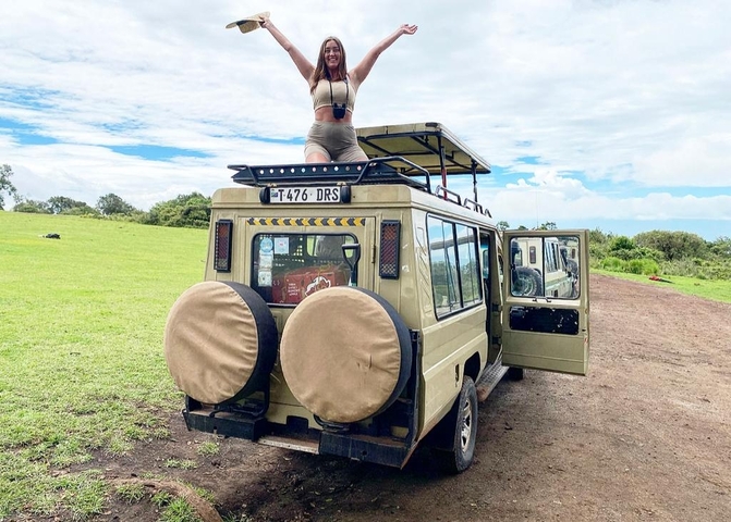 A person standing on a safari vehicle in a grassy landscape.