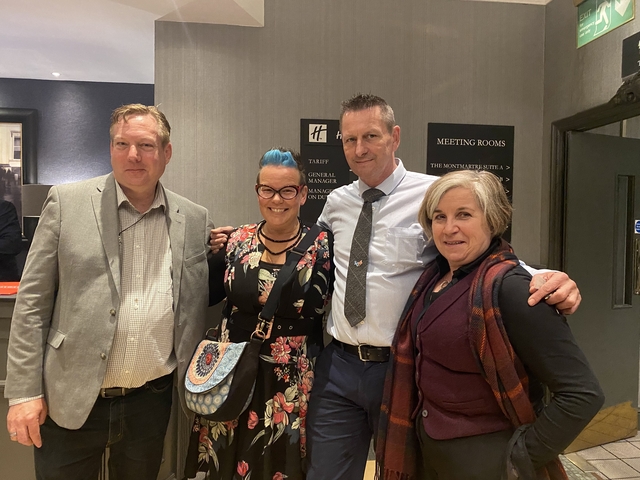       Four people posing inside a meeting room with signage.
  