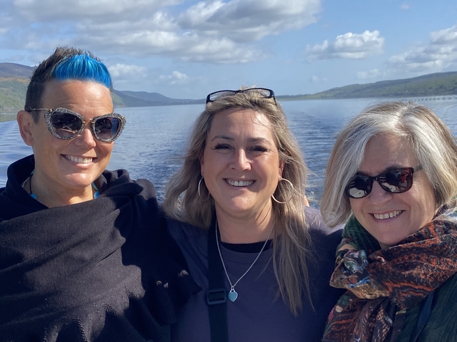       Three women smiling at the camera by a body of water.
  