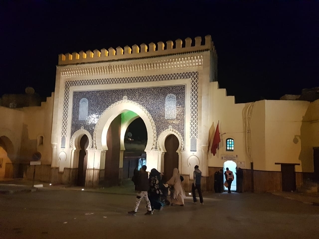       A Moroccan gate at night with people walking by.
  