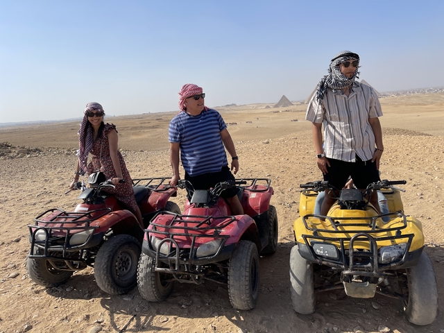 Three people on quad bikes with desert landscape and pyramids in the background.