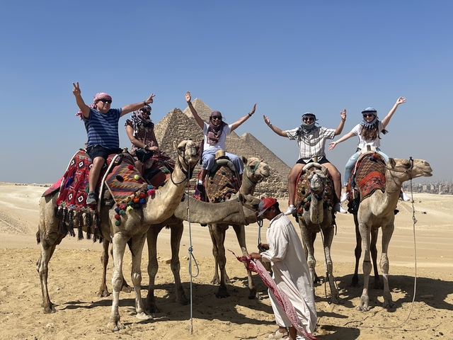       Group of people on camels cheering with pyramids in the background.
  