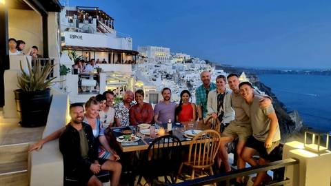       Group of people dining on a terrace with a view of white buildings.
  