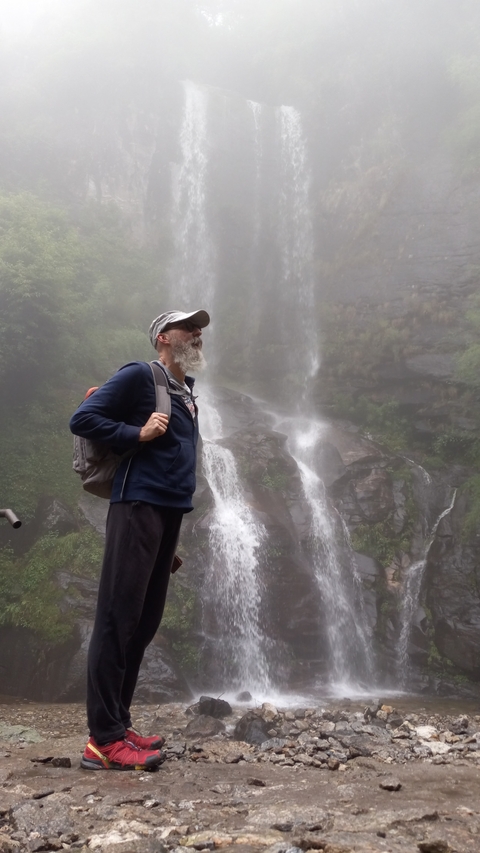 A man with a beard standing near a waterfall in a misty environment.