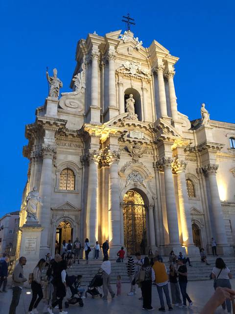       A large illuminated cathedral facade at night with crowds.
  