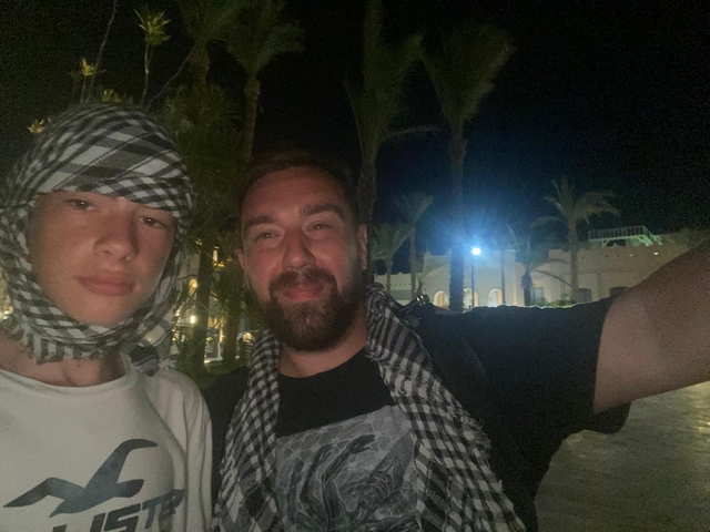 Two individuals taking a selfie with palm trees and buildings in the background.