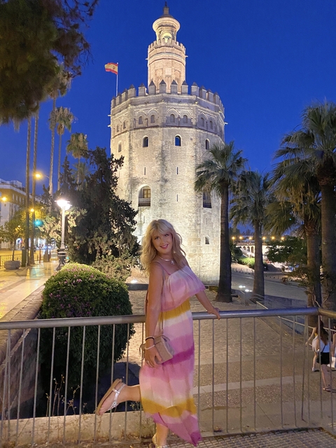       A woman in a colorful dress in front of a medieval tower with palm trees at night.
  
