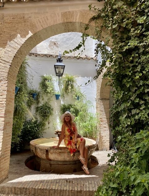       A woman in a floral dress and hat posing under an archway with potted plants.
  