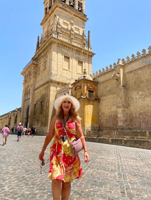       A woman in a dress and hat smiling in front of an old stone building.
  