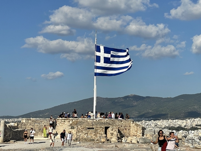       Greek flag waving with people gathered around a viewpoint.
  