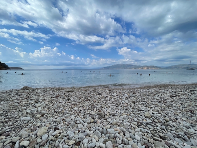       Pebble beach with swimmers in a calm sea under a partly cloudy sky.
  