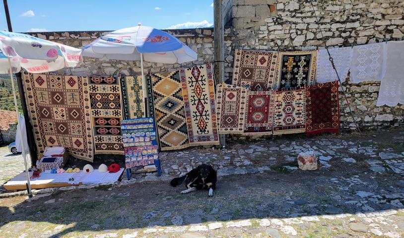       Colorful traditional woven rugs hung against a stone wall with a resting dog nearby.
  