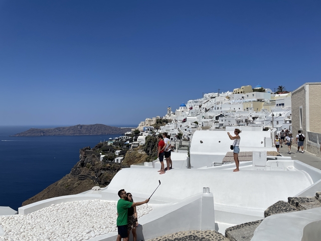       Tourists taking photos with a view of the sea and white buildings in Santorini.
  