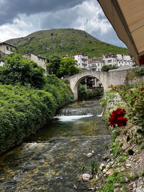       A stone bridge over a river with buildings and hills in the backdrop.
  