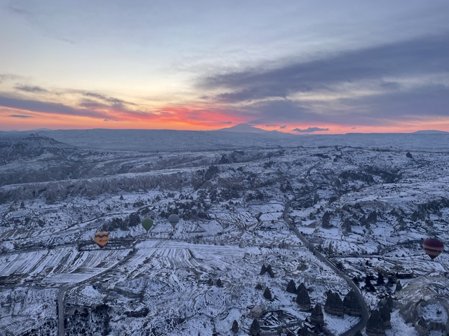 Scenic landscape of Cappadocia with hot air balloons and snowy mountains at sunset.