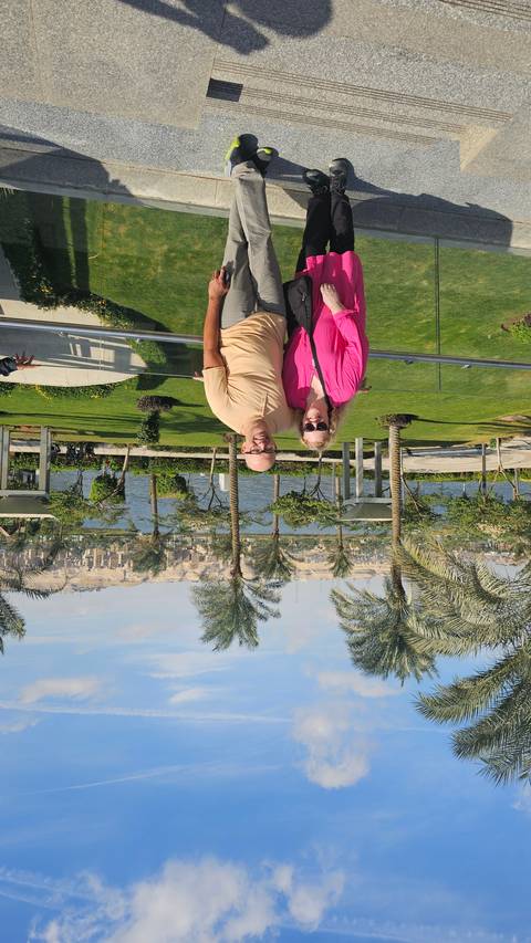       Two smiling people posing in a garden with palm trees and a city view in the background.
  