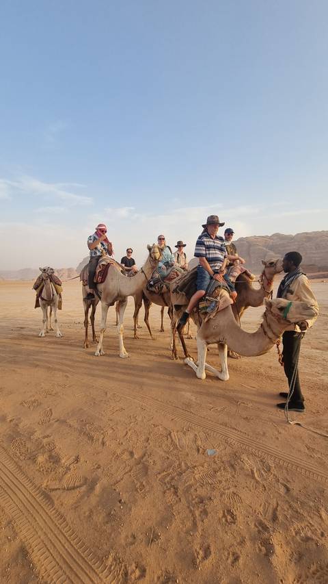       Tourists riding camels in a desert setting with mountains in the distance.
  