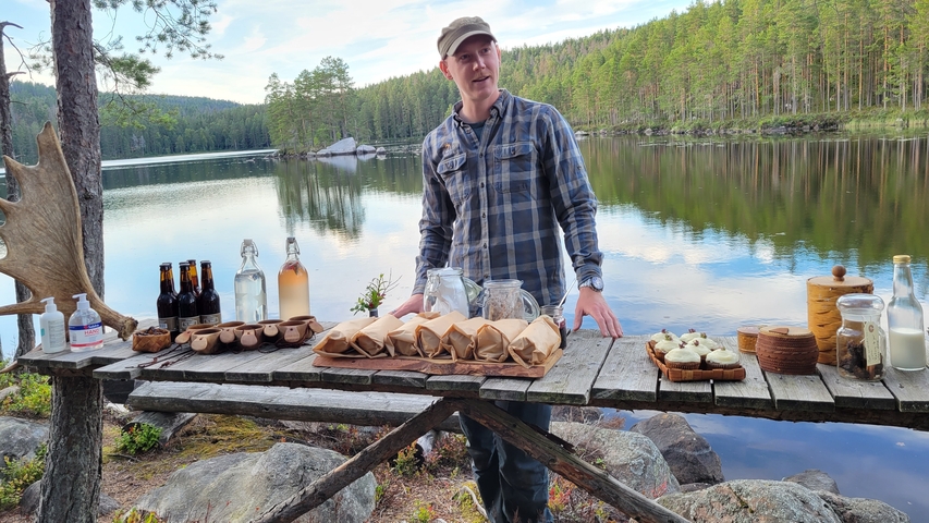 Man presenting a variety of drinks and snacks by a lake.