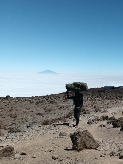       A person carrying a large bag on their head, walking in a barren landscape above the clouds.
  