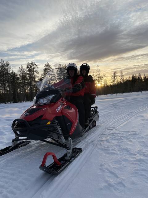 Two people on a snowmobile during sunset in a snowy forest.