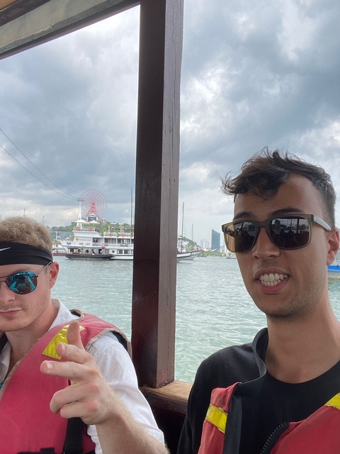 Two people on a boat with ferris wheel and ships in the background.