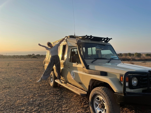       Person posing energetically with a safari vehicle in a savannah.
  