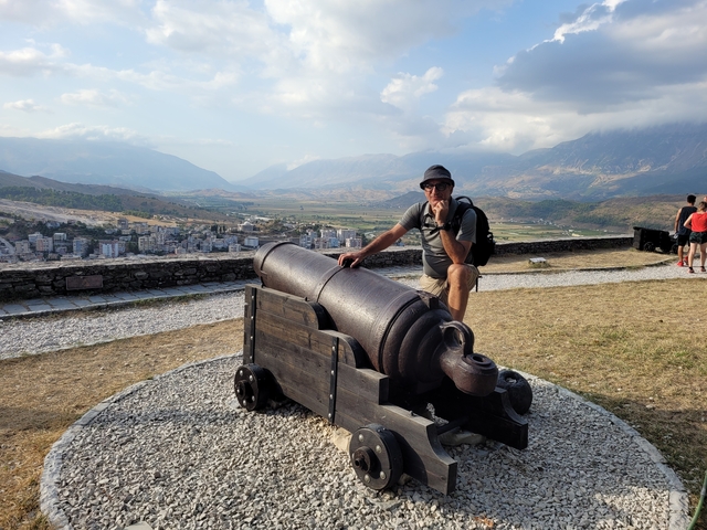 Person posing with an old cannon on a hill overlooking a town.