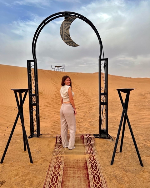 Woman standing in an elaborately decorated archway with sand dunes in the background.