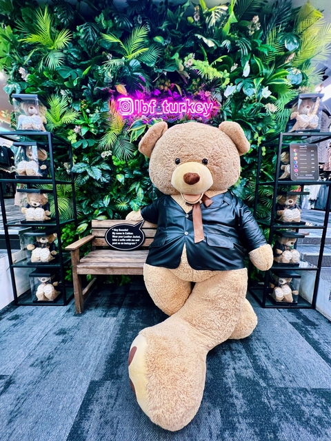       A large teddy bear wearing a leather jacket in a green setting.
  