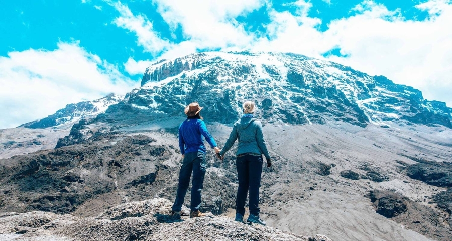 Two people holding hands stand on a rocky surface with Mount Kilimanjaro in the background.
