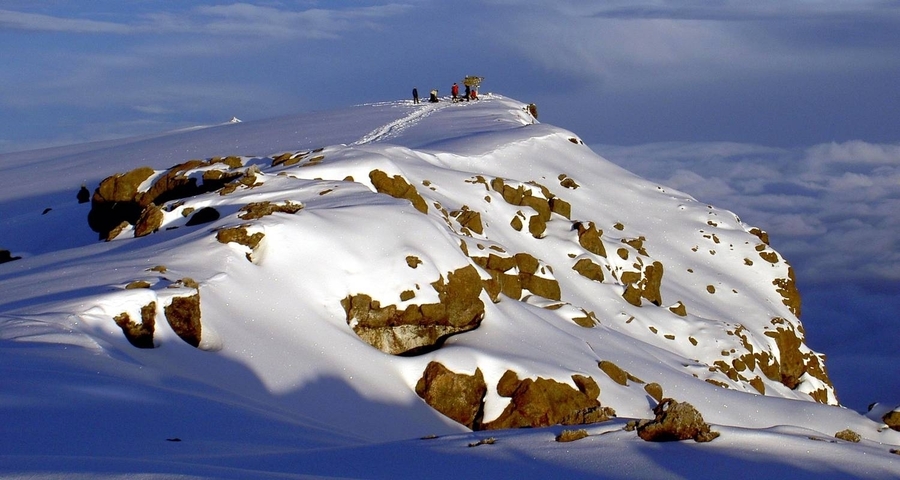 A snowy peak with a group of people walking on it under a cloudy sky.