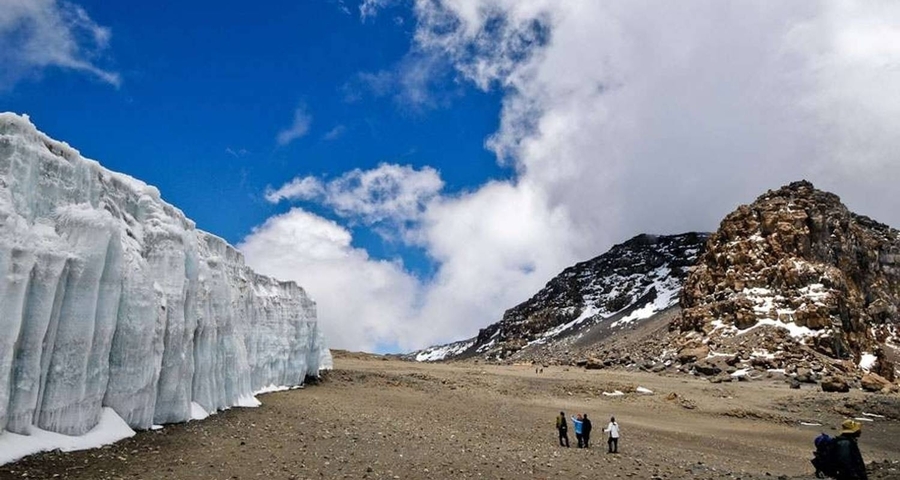 Glaciers and rocky formations under a blue sky, with people hiking.