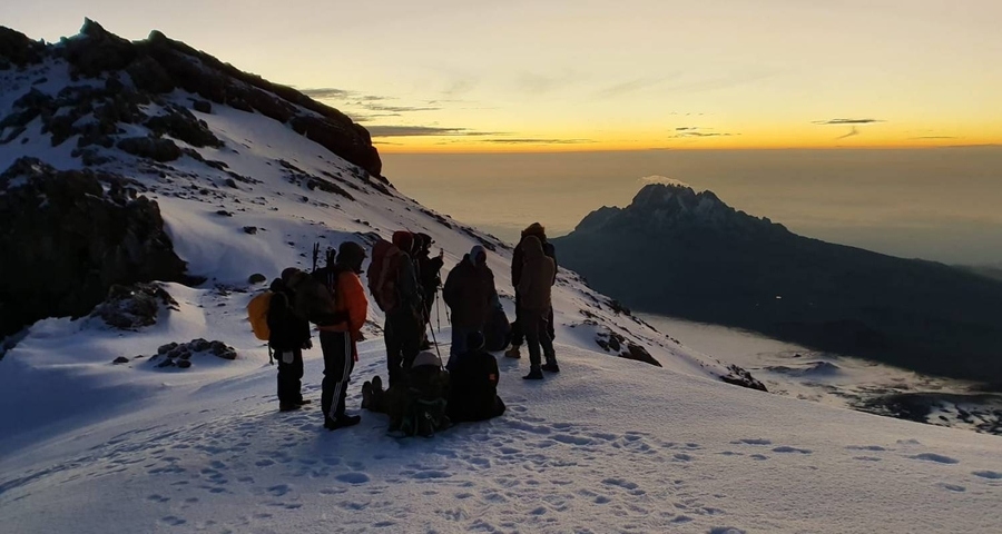 A group of hikers resting on a snowy mountain with a distant view of another peak at sunset.