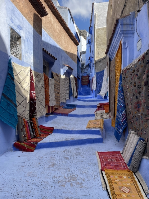       Narrow alley with colorful rugs displayed on blue walls.
  