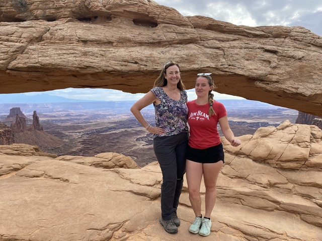       Two women standing under a natural rock arch with canyons.
  