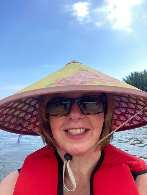 Person wearing sunglasses and traditional conical hat, smiling outdoors.