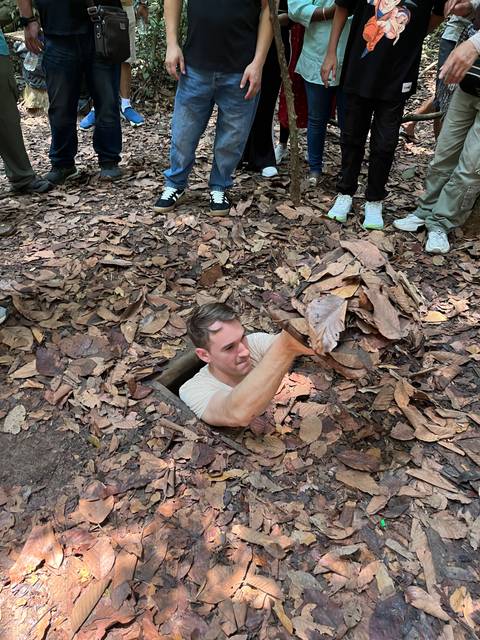 A person emerging from a small hole in the ground covered with dry leaves, with people standing nearby.