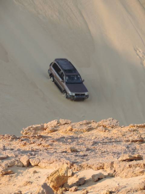 Car driving on desert sand with a rocky cliff.