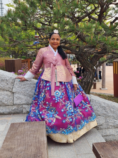 Woman in a colorful hanbok standing beside trees.