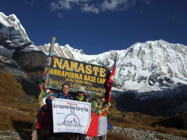       People at Annapurna Base Camp with sign and snowy mountains.
  