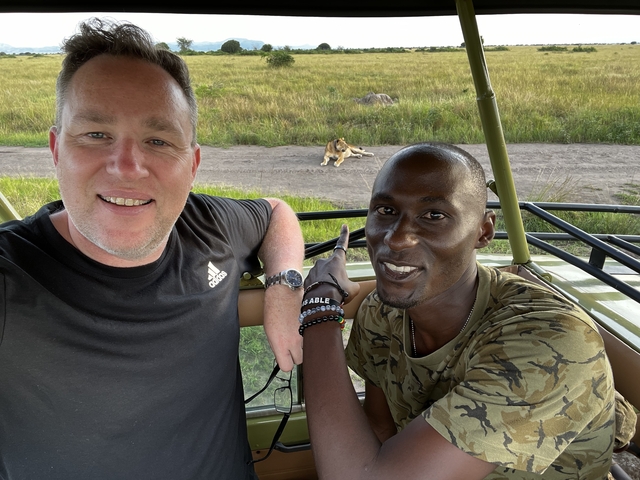       Two people in a safari vehicle with a lion resting in the background.
  