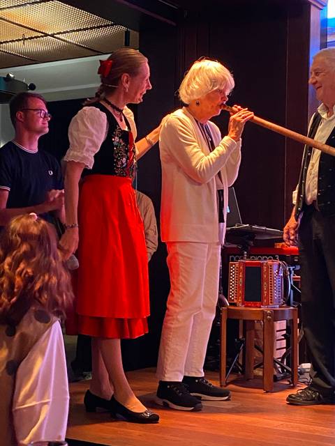       Elderly woman playing a long horn with others nearby.
  