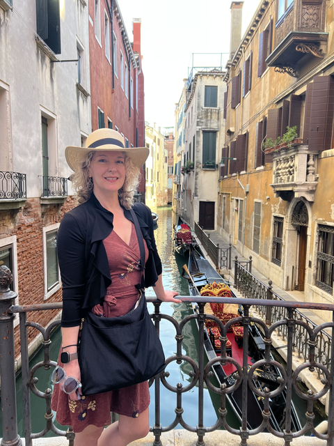 Woman wearing a hat standing on a bridge over a canal with gondola.