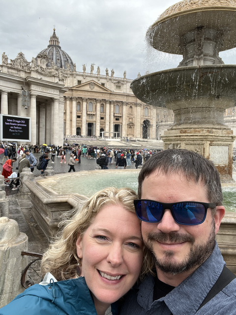 Close-up of two people with St. Peter's Basilica and fountain in the background.
