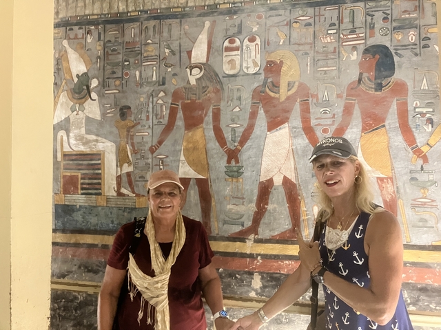 Two women posing in front of a mural featuring Egyptian-style art.