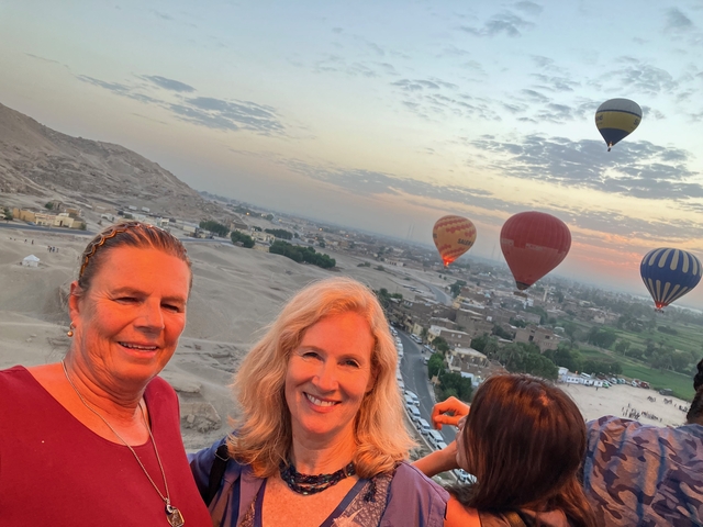 Two women smiling with hot air balloons in the background over a desert landscape.