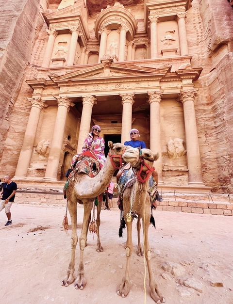 Two people riding camels in front of a rock-carved facade.