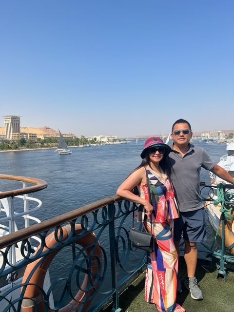       Two people posing on a boat with a river and buildings in the background.
  